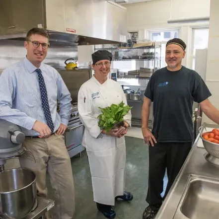 厨房里的三个用餐人员. 一个男人站在一碗西红柿旁边，一个女人拿着一捆蔬菜.